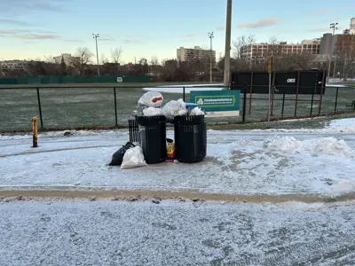 Garbage and recycling cans in a park overflowing with trash bags.