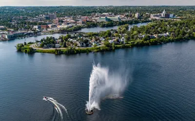 A large fountain shooting up into the air in the middle of a lake. A boat is navigating around the fountain. There are buildings sprawled out around the edge of the lake. 