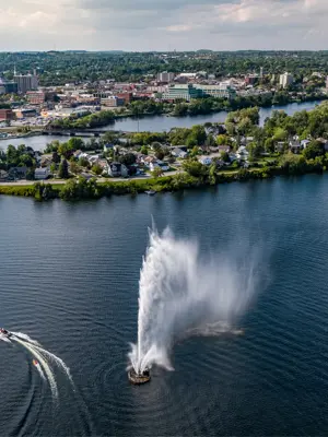 A large fountain shooting up into the air in the middle of a lake. A boat is navigating around the fountain. There are buildings sprawled out around the edge of the lake. 